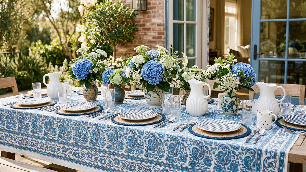 Elegant outdoor dining table setting with blue and white tablecloth, floral centerpieces, and cutlery.