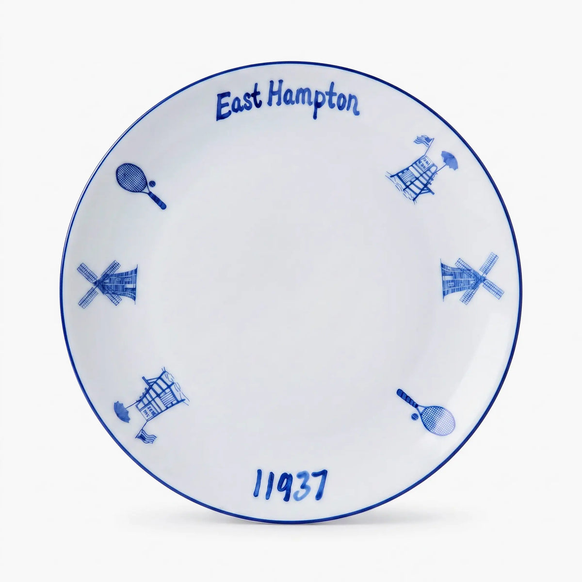 A white ceramic commemorative plate featuring blue hand-painted details, including the text 'East Hampton' and '11937', along with windmill and tennis racket motifs, set against a clean white background.