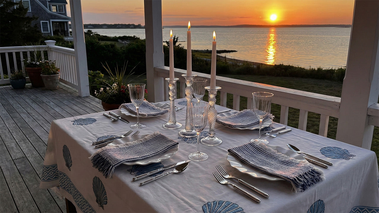 An outdoor dinner table on a white wooden porch overlooking a bay or ocean at sunset.