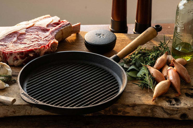 Grill pan on a wooden surface with raw meat, herbs, and bottles in the background