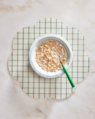 White bowl with cereal on a green and white checkered placemat on a marble surface