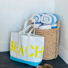 Beach bag with 'BEACH' text, rolled towels, and sandals on a dark surface.