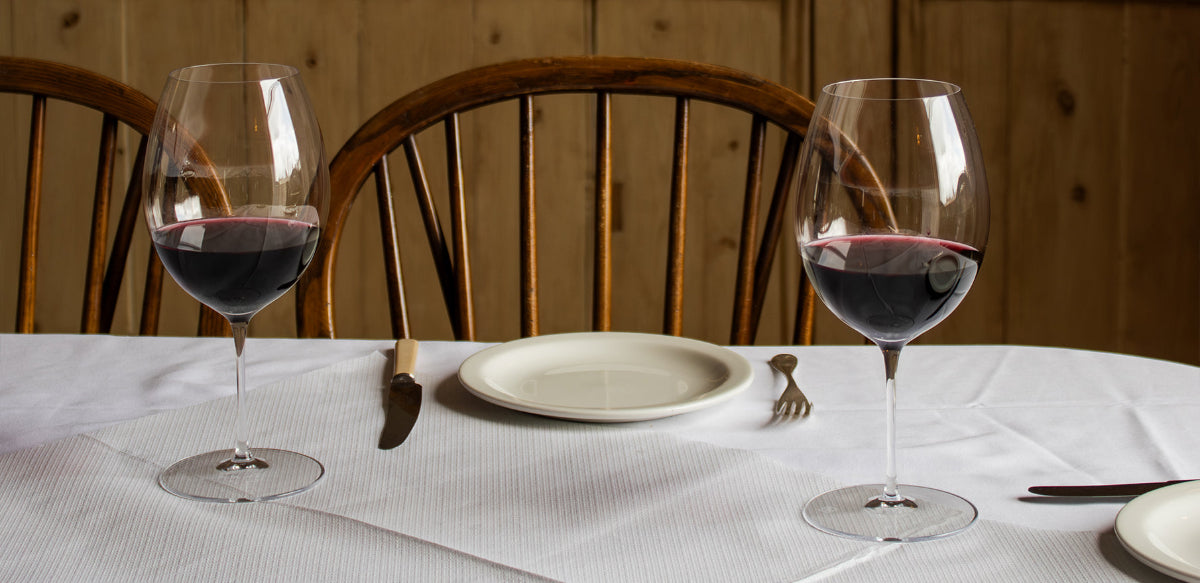 Two wine glasses with red wine on a table set for two, with a wooden chair in the background.