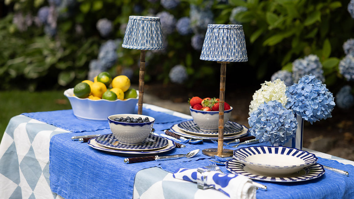 An outdoor brunch table setting featuring blue and white patterned linens, hydrangea arrangements, and small cordless lamps with blue patterned shades.