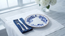 A white ceramic plate with a blue floral wreath pattern, styled on a white table runner with a navy blue napkin, silver cutlery, and white flowers in the background.
