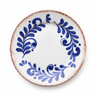 A round white ceramic plate with a rustic, hand-painted blue floral pattern and a textured, reddish-brown rim, shown from a top-down perspective on a white background.