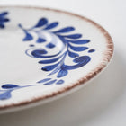 A close-up of a white ceramic plate featuring a blue hand-painted floral pattern and a rustic, slightly uneven brown rim.