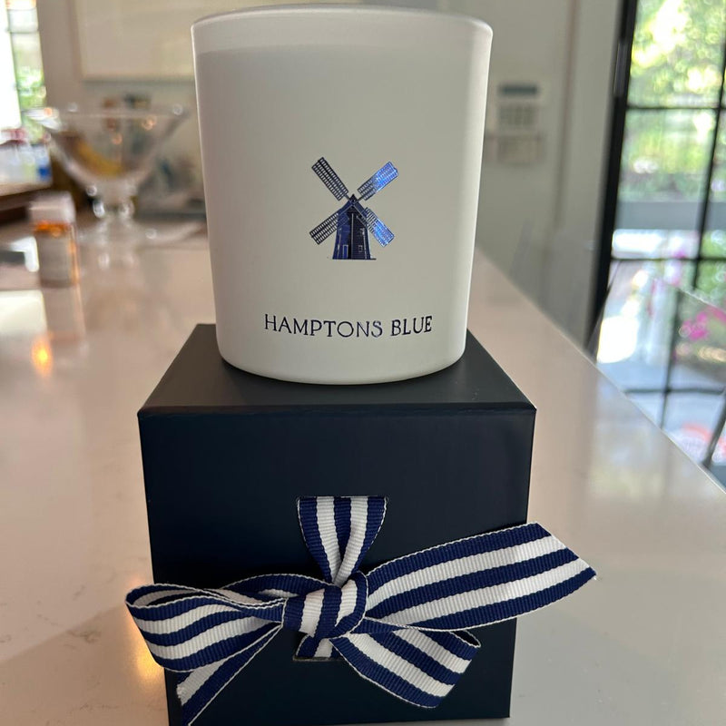 White candle with 'Hamptons Blue' branding on a navy box with a striped ribbon.