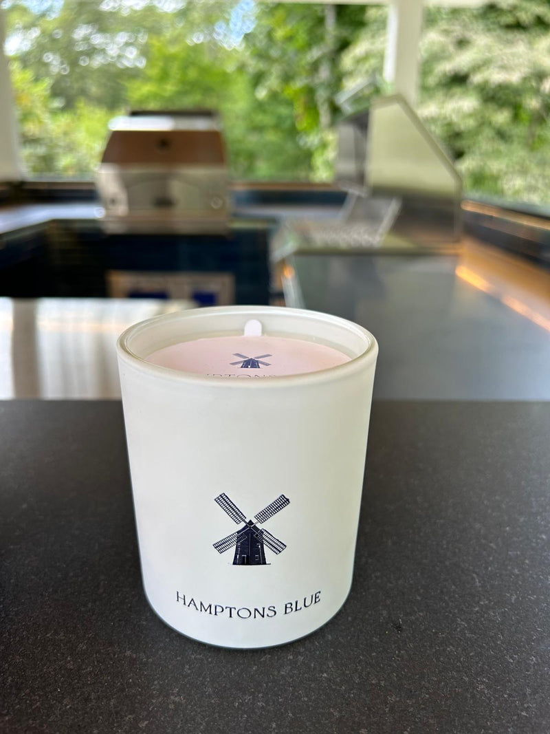 A white candle from the brand "HAMPTONS BLUE" featuring a windmill logo.