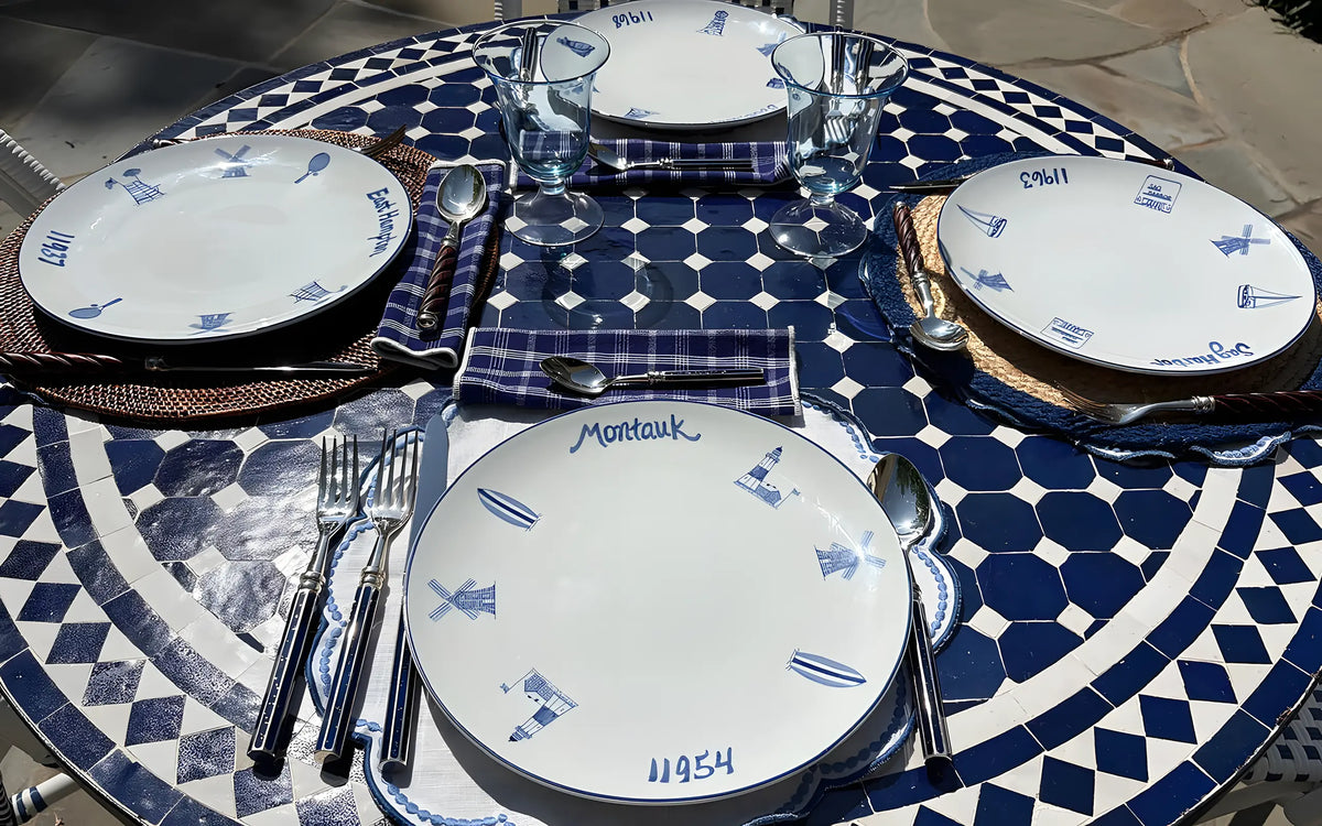 A round dining table set with four place settings featuring white plates with blue illustrations, blue-and-white patterned table linens, silver cutlery, and clear drinking glasses.