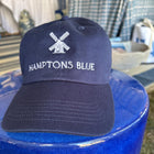 Navy blue cap with 'Hamptons Blue' text and logo on a blue surface.