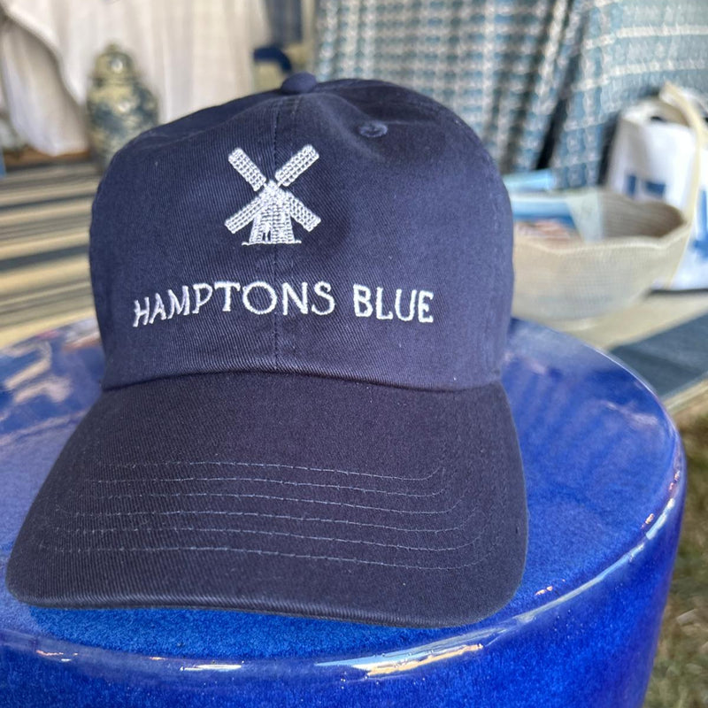 Navy blue cap with 'Hamptons Blue' text and logo on a blue surface.