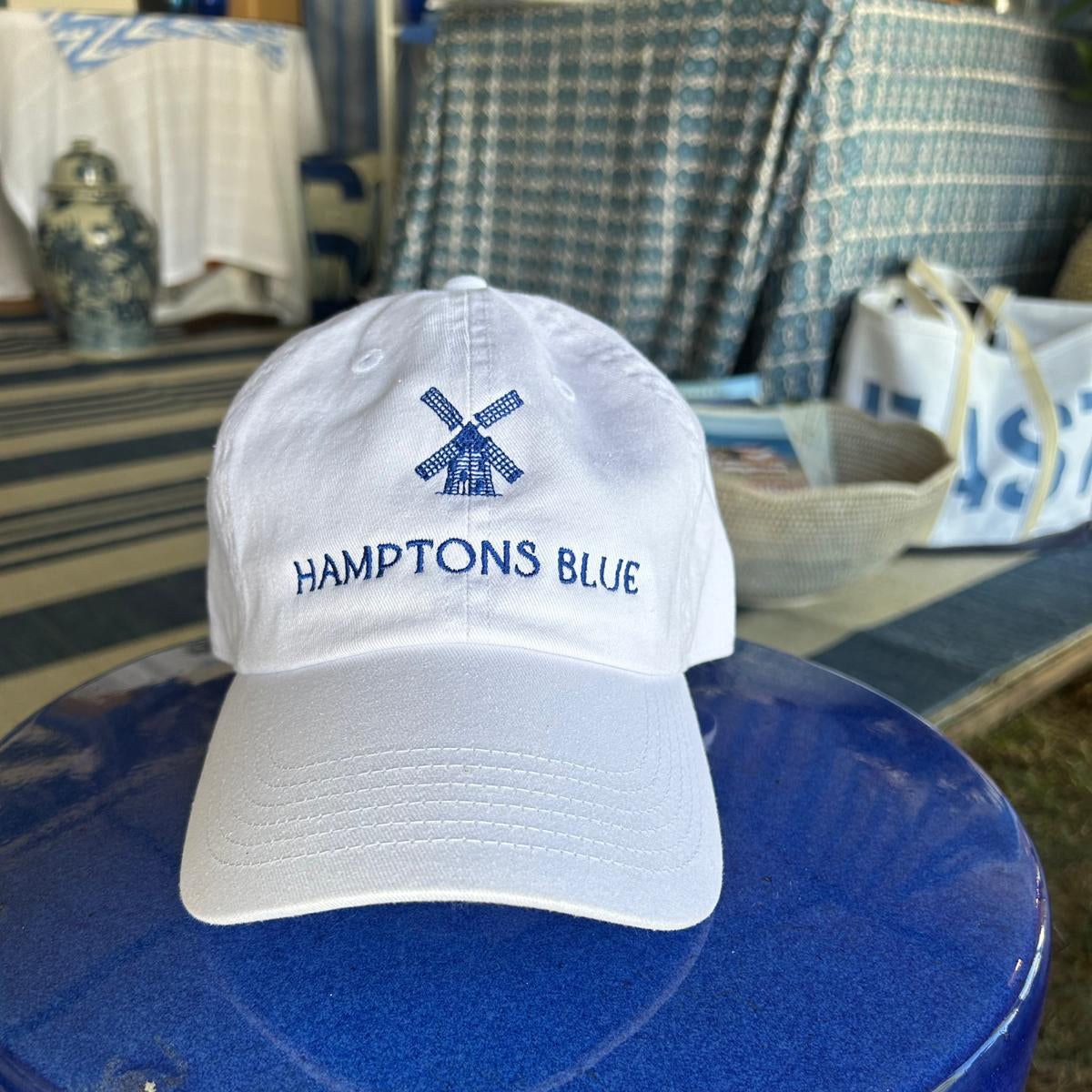 White cap with 'Hamptons Blue' logo on a blue stool outdoors.