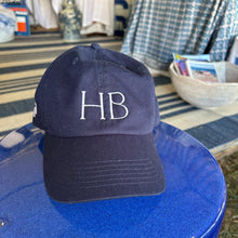 Navy blue cap with 'HB' initials on a blue surface, with a blurred background of a store interior.