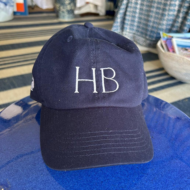 Navy blue cap with 'HB' initials on a blue surface, with a blurred background of a store interior.