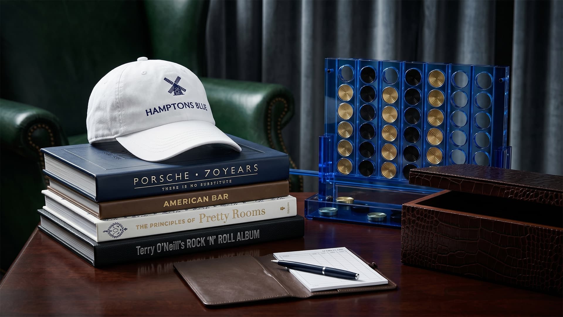 Stack of books with a cap on top, next to a blue dice set and leather-bound notebook on a wooden surface.