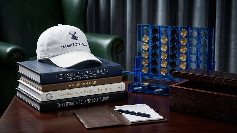 Stack of books with a cap on top, next to a blue dice set and leather-bound notebook on a wooden surface.