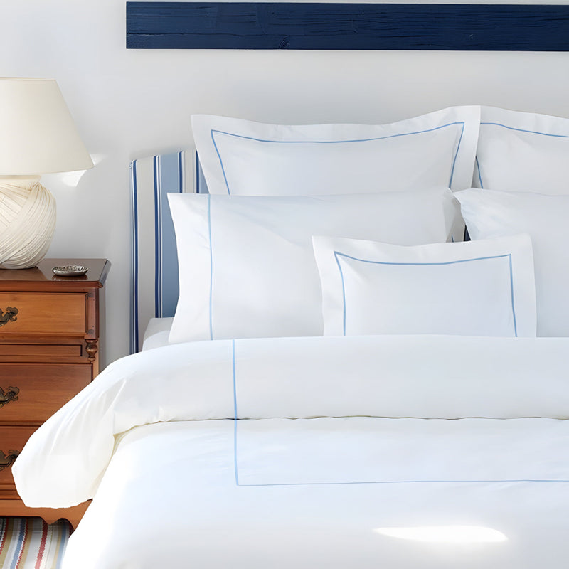 White bedding set with blue piping on a bed with a wooden nightstand and lamp.