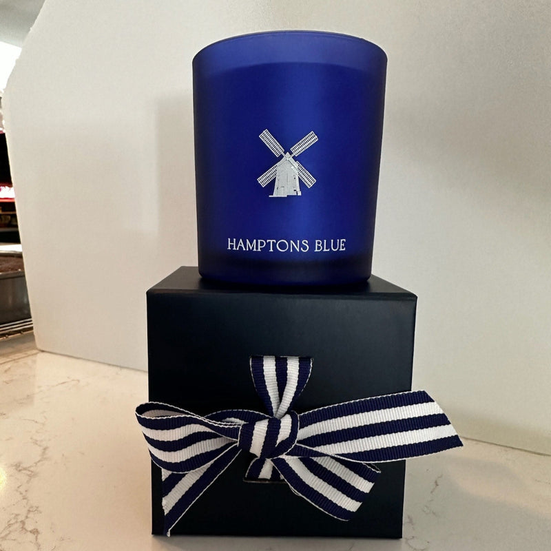 A blue candle from the brand "HAMPTONS BLUE" featuring a windmill logo  is presented on a dark blue gift box with a striped ribbon.