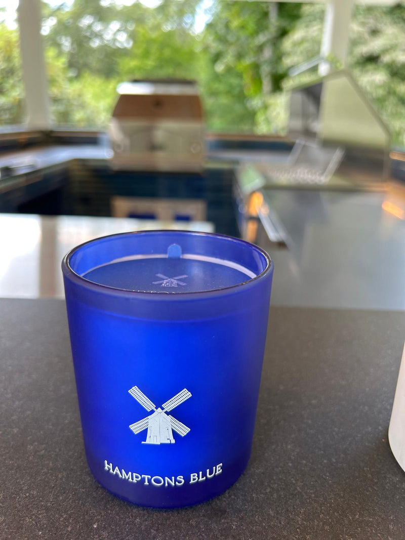 A blue candle from the brand "HAMPTONS BLUE" featuring a windmill logo.