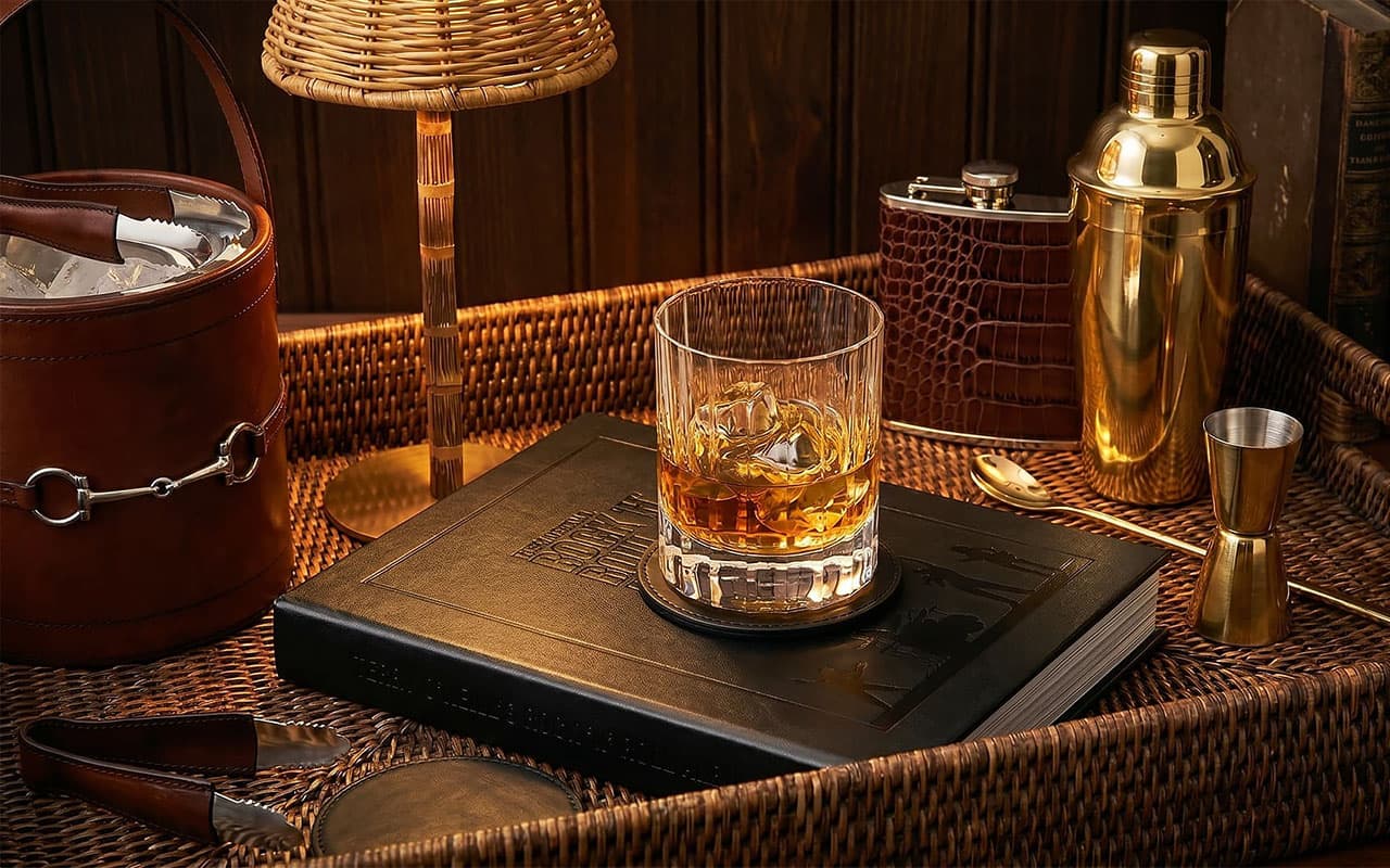 Glass of whiskey on a wooden coaster with bar tools in the background