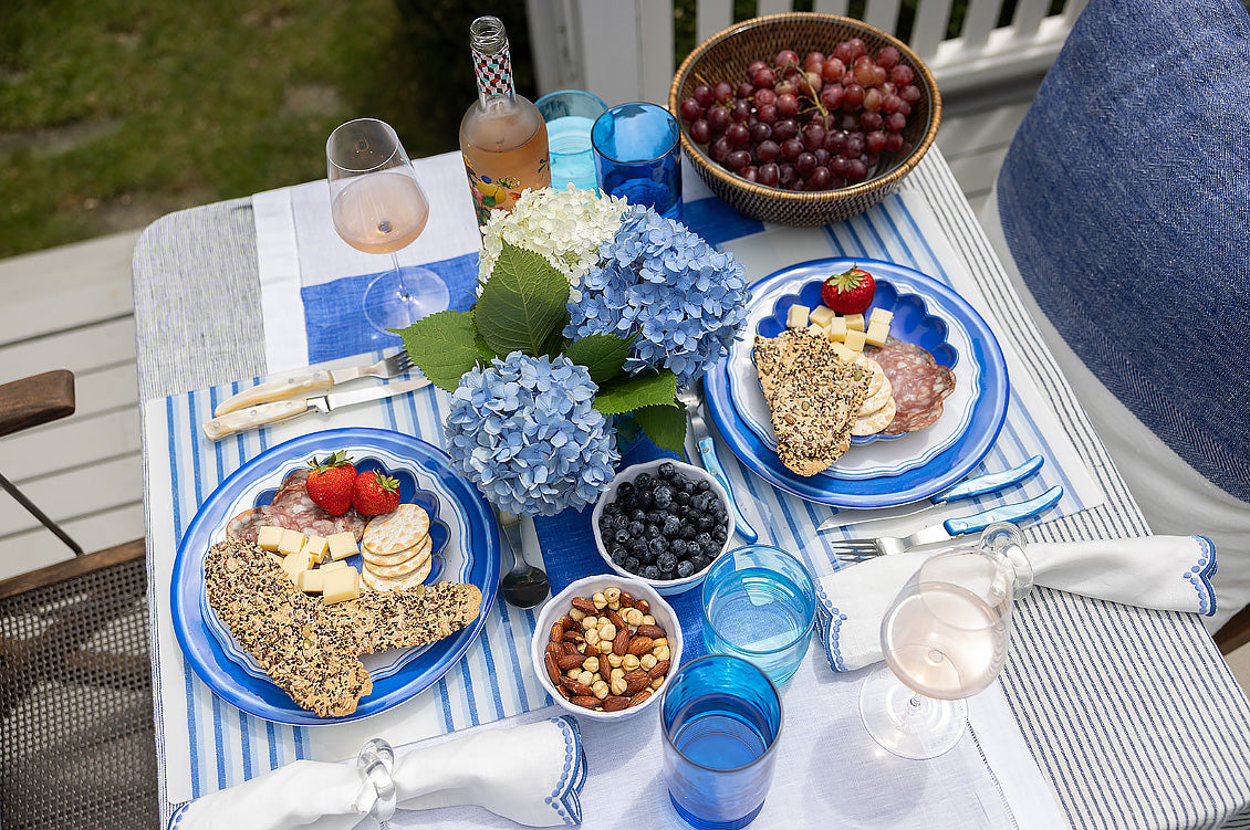 Outdoor table setting with plates of food, drinks, and flowers on a striped tablecloth.