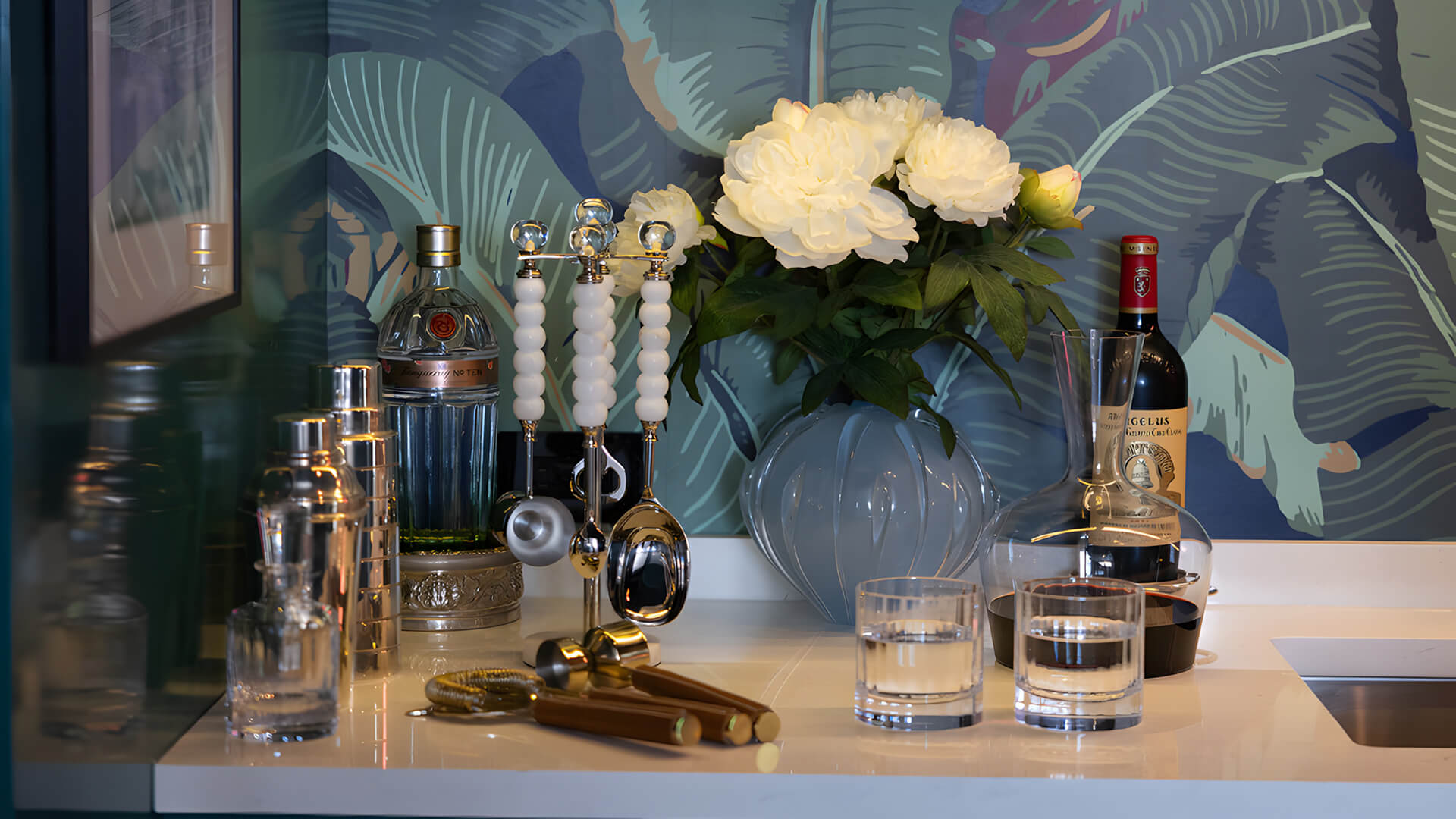 Alcohol bottles, glasses, and a vase with flowers on a bar counter against a decorative wall.