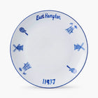 A white ceramic commemorative plate featuring blue hand-painted details, including the text 'East Hampton' and '11937', along with windmill and tennis racket motifs, set against a clean white background.