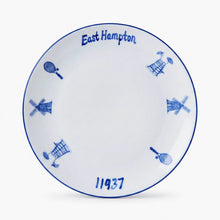 A white ceramic commemorative plate featuring blue hand-painted details, including the text 'East Hampton' and '11937', along with windmill and tennis racket motifs, set against a clean white background.