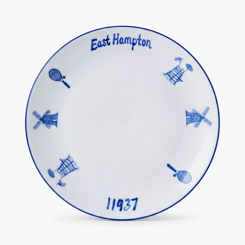 A white ceramic commemorative plate featuring blue hand-painted details, including the text 'East Hampton' and '11937', along with windmill and tennis racket motifs, set against a clean white background.