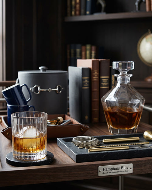 Whiskey glass with ice, decanter, and books on a wooden table.