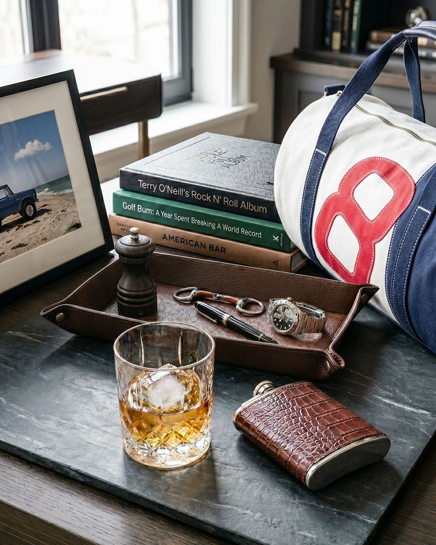 Glass of whiskey with ice, leather flask, and duffel bag on a table.