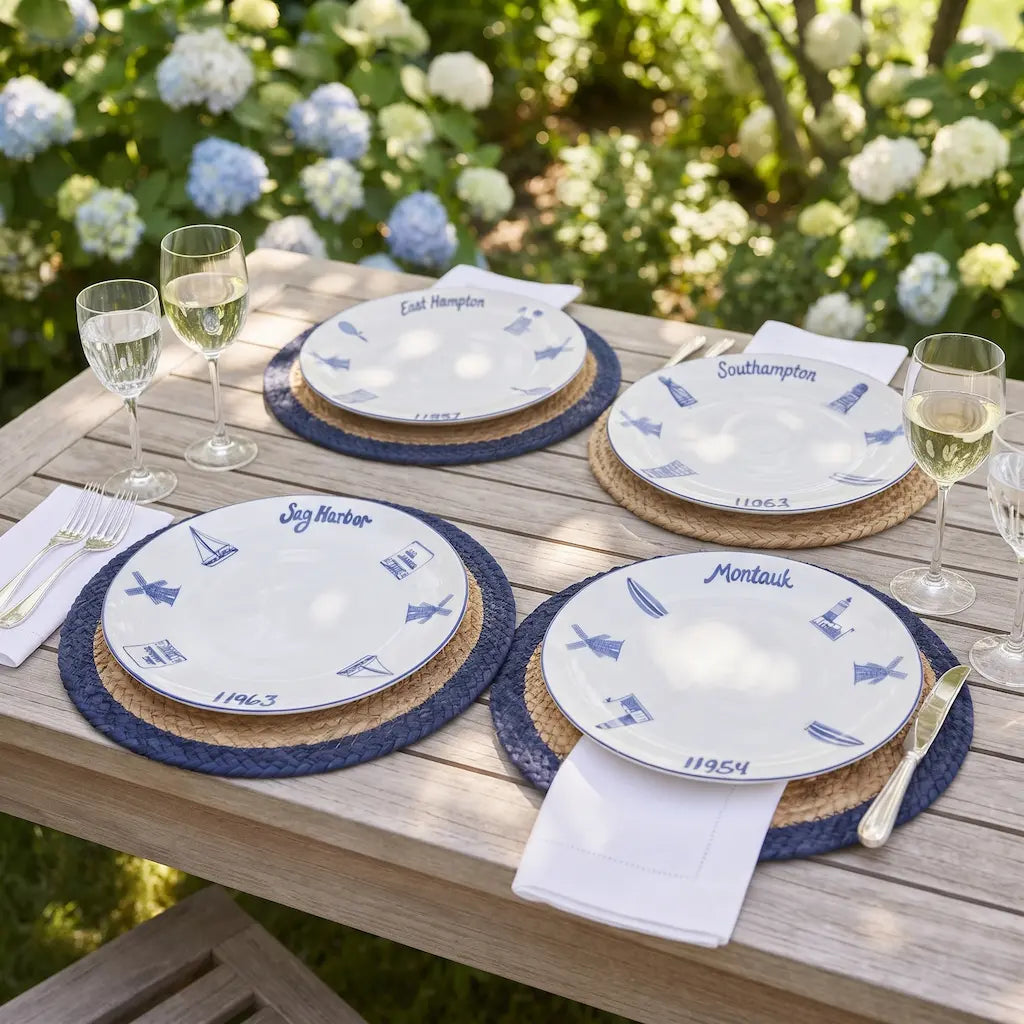 Outdoor dining table setting with white plates printed with blue nautical motifs and names, layered on woven placemats with white napkins, silverware, and wine glasses, with blurred greenery and flowers in the background.
