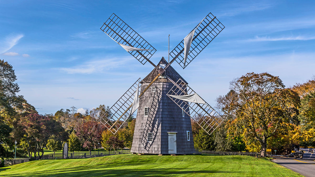 A tall gray wooden windmill with large metal blades stands on a green lawn, surrounded by trees with autumn foliage under a bright blue sky.