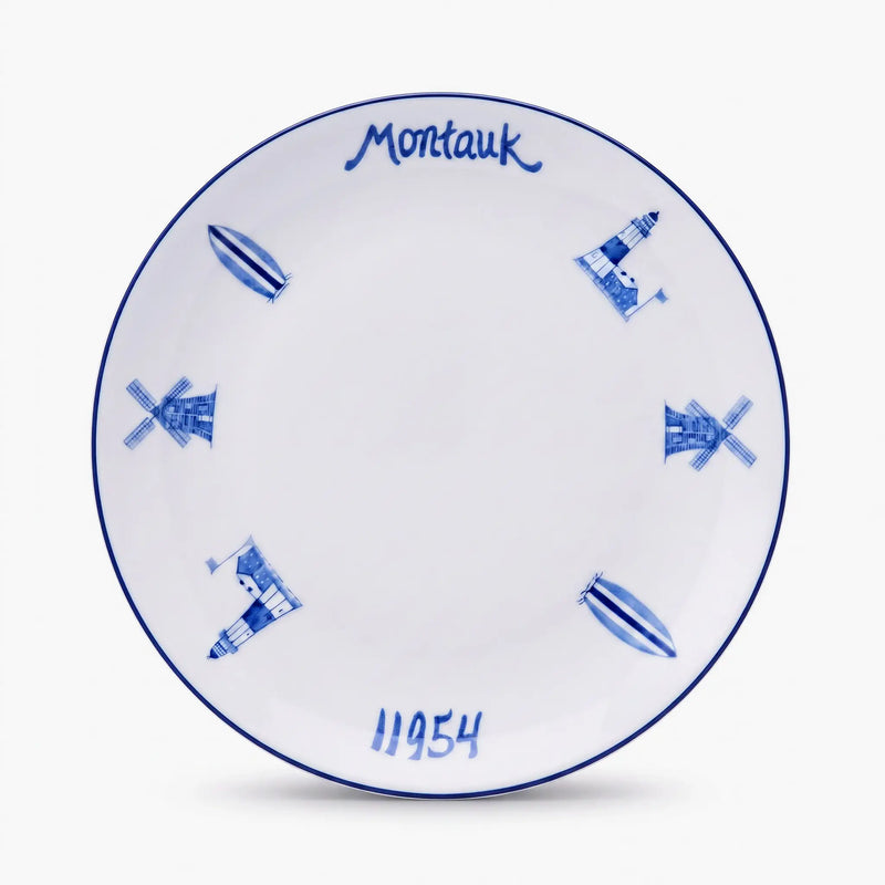 A white ceramic plate with blue hand-painted illustrations of lighthouses, windmills, and boats, featuring the text 'Montauk' at the top and '11954' at the bottom, shown on a light gray background.