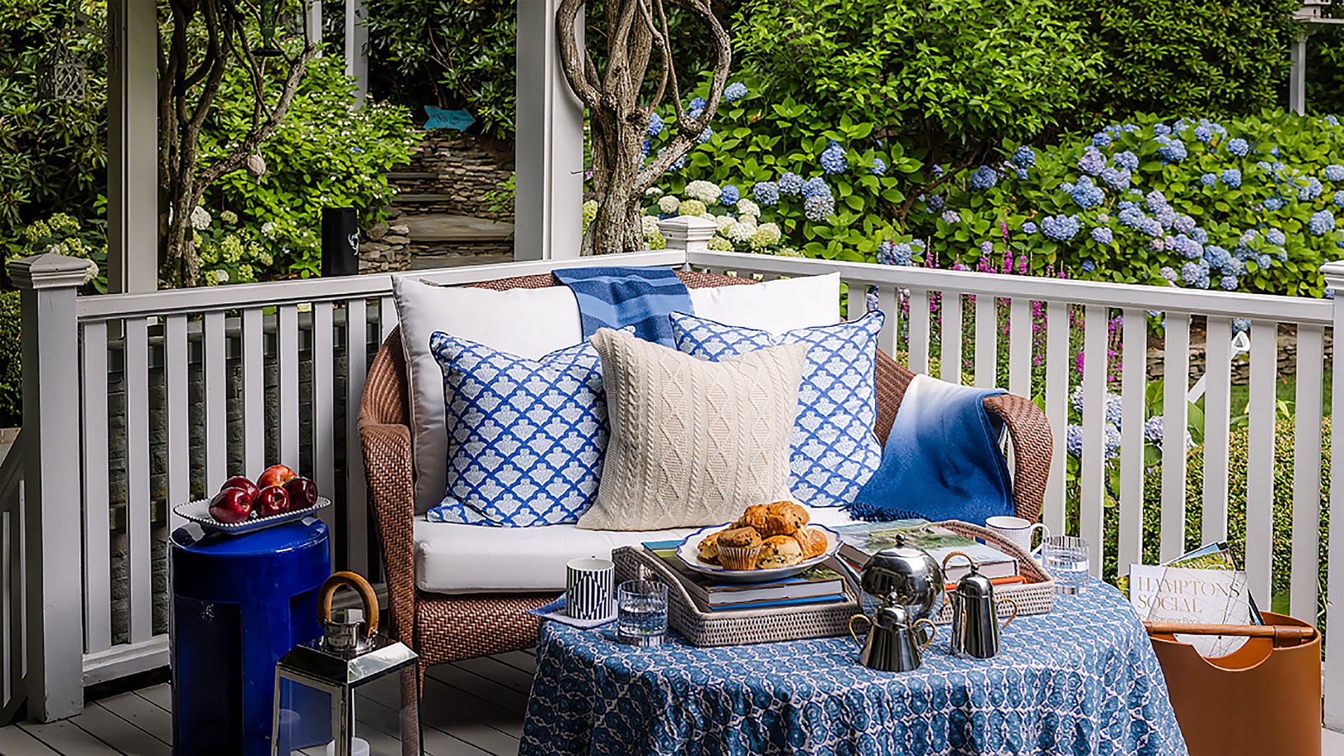 Outdoor patio with wicker chair, cushions, and a table set for a meal with a garden view.