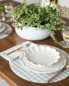 Set of white ceramic plates with on a green and white checked placemat on a wooden table with a plant in the background.