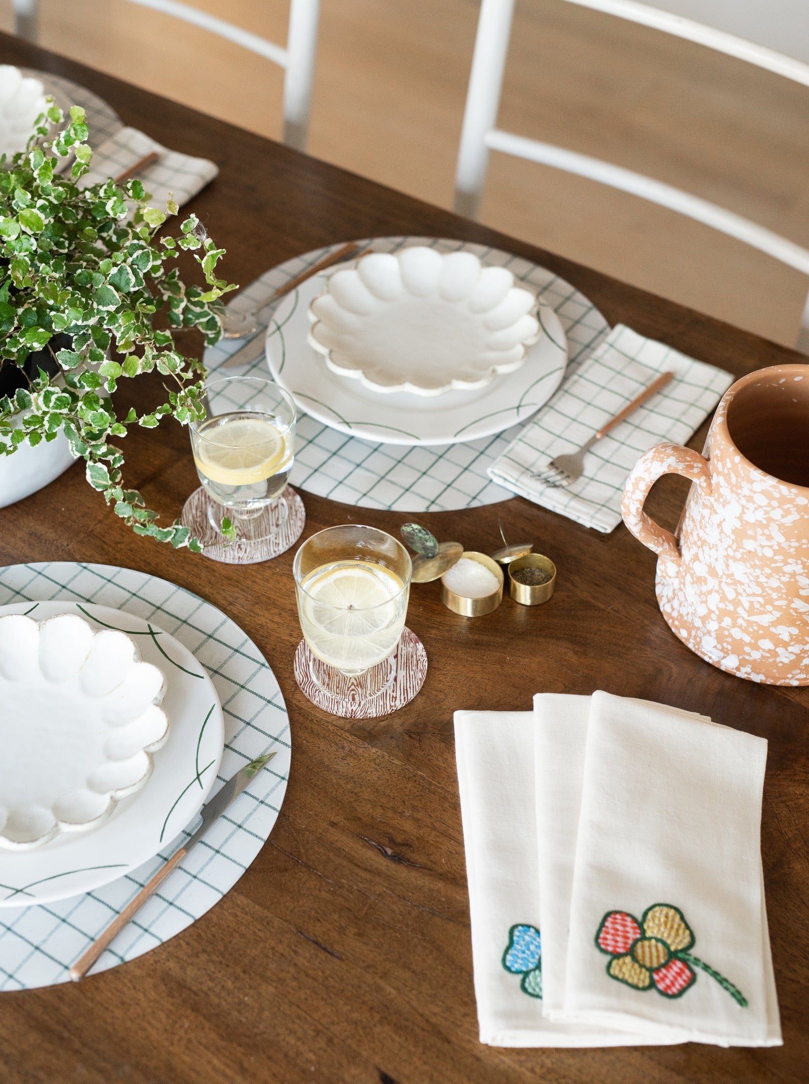 Dining table setting with plates, glasses, placemats and embroidered napkins on a wooden table.