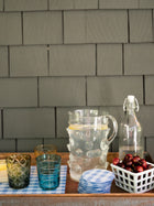 Outdoor setting with glasses, a pitcher, a stack of coasters and a bottle on a table against a shingled wall.