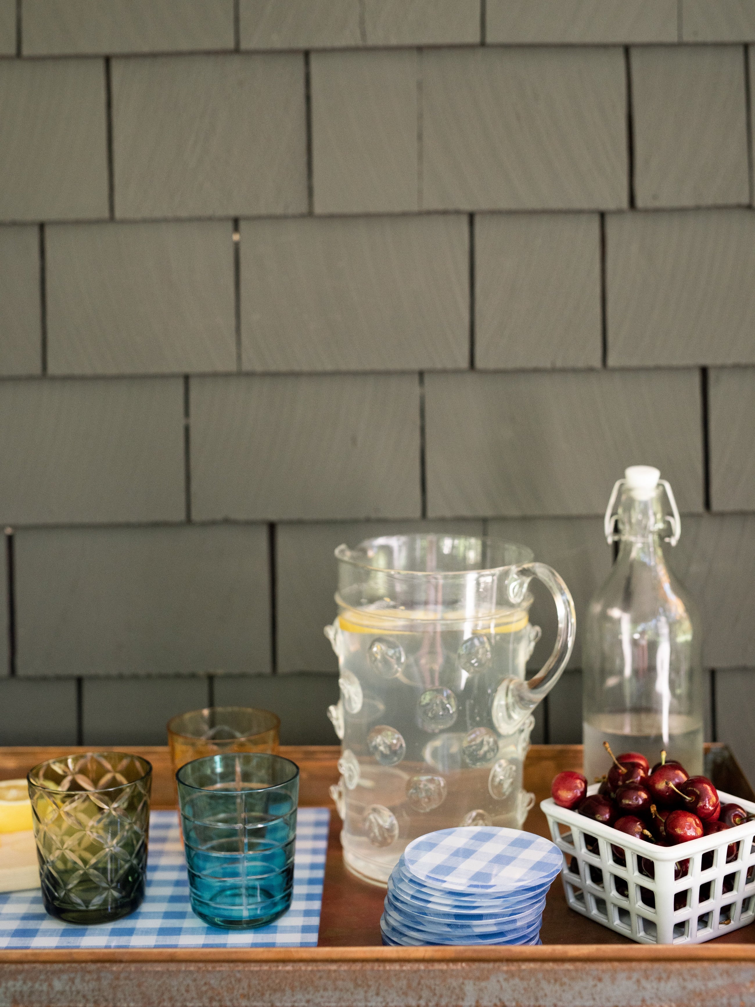 Outdoor setting with glasses, a pitcher, a stack of coasters and a bottle on a table against a shingled wall.