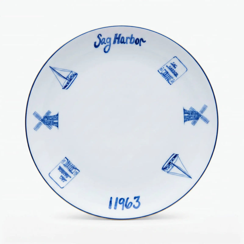 A white ceramic plate with blue hand-painted illustrations of sailboats, windmills, and stamps, featuring the text 'Sag Harbor' and '11963' on a clean white background.